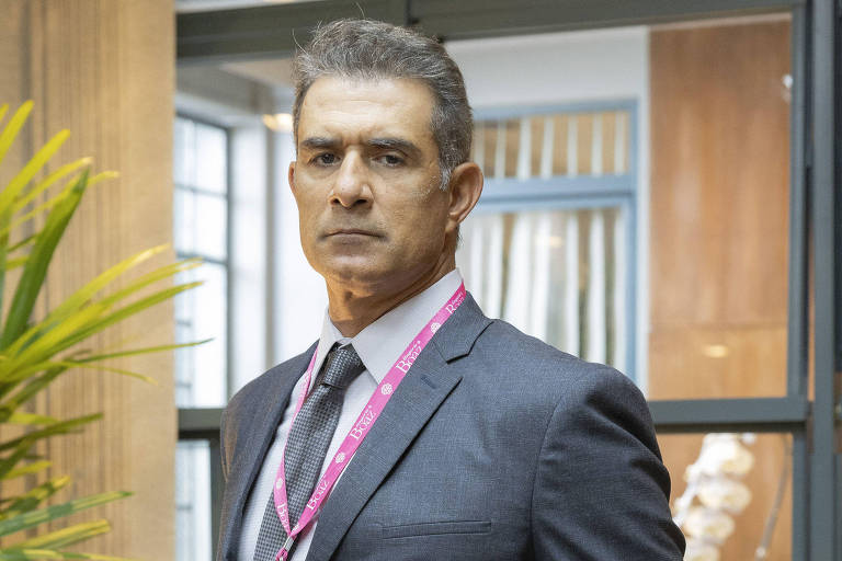 Um homem em traje formal, usando um terno escuro e uma gravata. Ele está em pé, com uma expressão séria, em um ambiente interno que parece ser um escritório. Ao fundo, há uma planta decorativa e uma cadeira de couro. O homem tem um crachá pendurado no pescoço e uma mão no bolso.