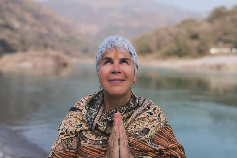 Uma mulher com cabelo curto e grisalho está em pé à beira de um rio, com as mãos unidas em um gesto de oração. Ela usa um xale colorido e olha para cima, em direção ao céu. Ao fundo, há montanhas e uma vegetação densa, com um céu claro e azul.