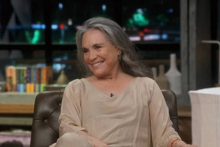 Regina Duarte, uma mulher com cabelos grisalhos e longos está sentada em um sofá em um estúdio. Ela usa uma blusa clara e sorri enquanto conversa. Ao fundo, há prateleiras com livros e uma iluminação suave, criando um ambiente acolhedor.