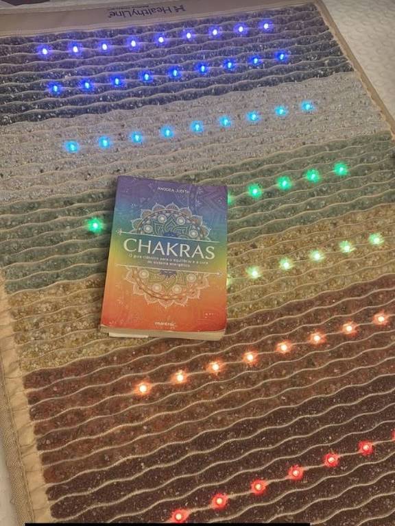 A imagem mostra um livro intitulado "Chakras" posicionado sobre um tapete com luzes coloridas. O tapete possui uma textura ondulada e as luzes estão dispostas em linhas horizontais, com cores variando entre verde, azul, vermelho e amarelo. A parte inferior da imagem contém um texto que menciona a sincronicidade do universo e a intenção de meditar.
