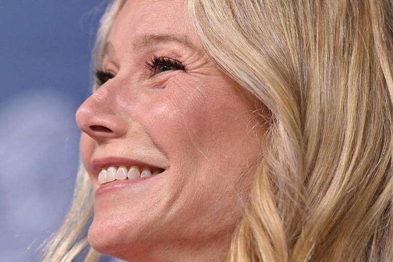 A imagem mostra um close de uma mulher com cabelo loiro e ondulado, sorrindo. Ela tem uma expressão alegre e está olhando ligeiramente para cima. O fundo é desfocado, com tons azuis e círculos que sugerem um evento ou conferência.