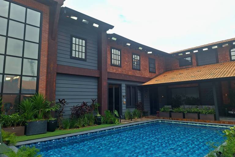 A imagem mostra uma casa moderna com uma piscina em primeiro plano. A casa possui paredes de madeira e uma cobertura de telhas. Há grandes janelas com molduras pretas e plantas ao redor da piscina. O céu está parcialmente nublado, e a área externa é bem iluminada.