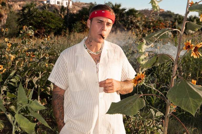 Um homem está posando em um campo de girassóis. Ele usa uma camisa listrada branca e calças brancas, além de um lenço vermelho na cabeça. O homem segura um cigarro e está cercado por plantas altas de girassóis. Ao fundo, há uma construção em uma colina e palmeiras.
