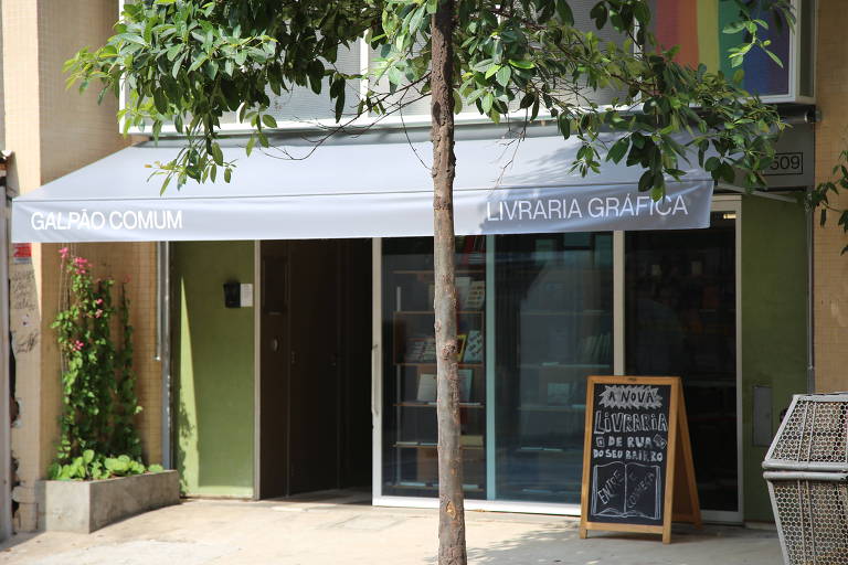 A imagem mostra a fachada de uma livraria gráfica chamada 'Livraria Gráfica', localizada em um espaço com toldo cinza. O toldo possui o nome da livraria em letras brancas. À esquerda, há uma porta de entrada e, ao lado, uma lousa de madeira com informações sobre a livraria. O ambiente é cercado por uma planta e a parede ao fundo é de cor verde. O dia parece ensolarado.