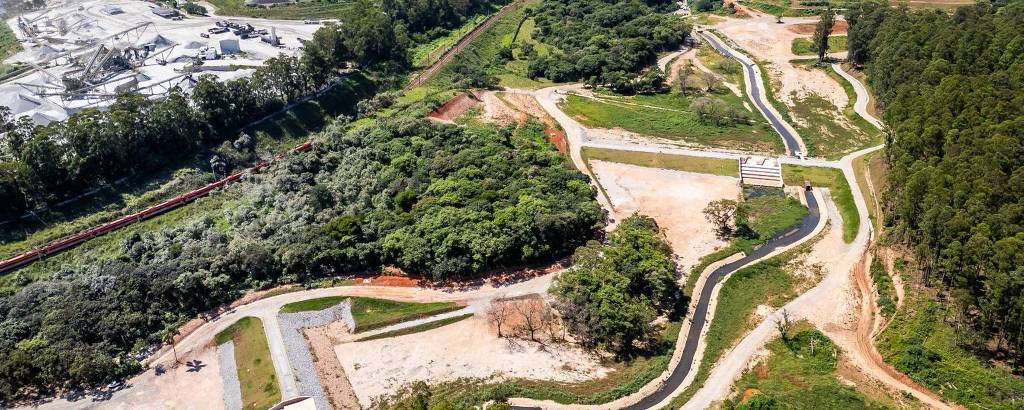 A imagem mostra uma vista aérea de uma área que combina urbanização e natureza. À esquerda, há uma construção em andamento, possivelmente um empreendimento urbano, enquanto à direita, observa-se uma área verde com árvores e um curso d'água sinuoso. Ao fundo, é visível uma cidade com várias edificações e montanhas ao longe.