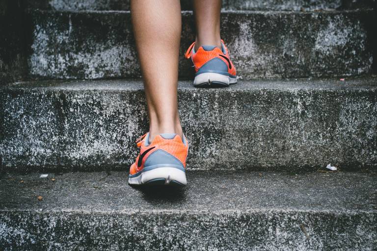 A imagem mostra uma pessoa subindo escadas. A parte inferior das pernas é visível, e a pessoa está usando tênis de corrida com detalhes em laranja e cinza. As escadas são de concreto e apresentam uma superfície desgastada.