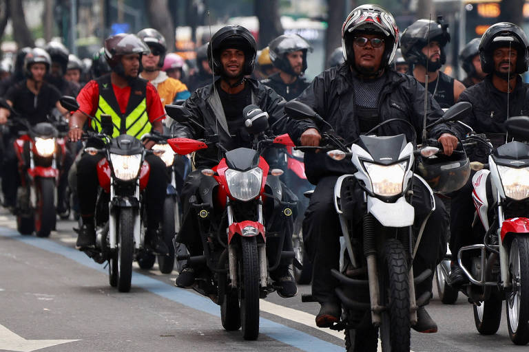 A imagem mostra um grupo de motociclistas em uma rua, todos usando capacetes. Alguns motociclistas estão vestidos com roupas escuras, enquanto outros usam coletes de segurança de cores vibrantes. Eles estão alinhados em uma faixa de moto, com várias motos de diferentes modelos e cores, predominantemente vermelhas e pretas. O ambiente parece urbano, com prédios ao fundo e um clima nublado.