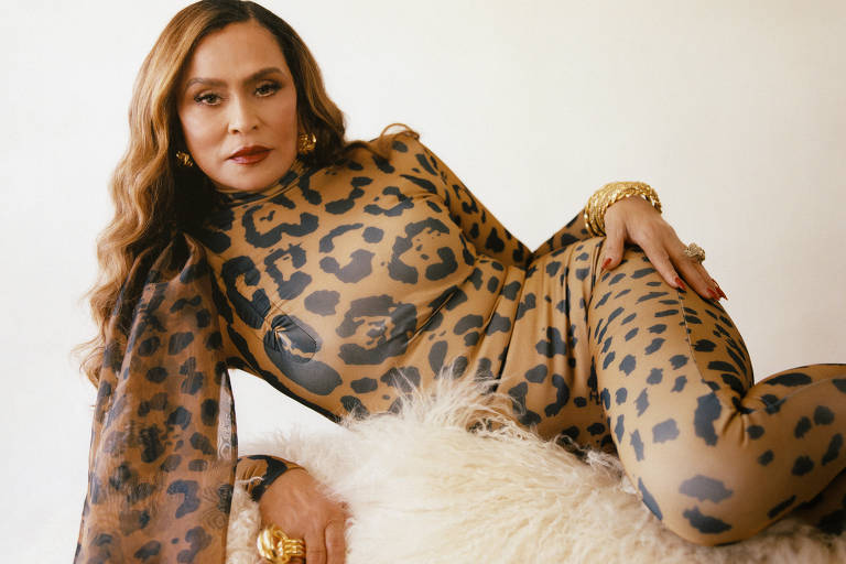 A imagem mostra uma mulher deitada sobre um fundo claro, vestindo um traje justo com estampa de onça. Ela tem cabelos longos e ondulados, e está usando acessórios dourados, incluindo anéis e pulseiras. A pose é relaxada, com uma mão apoiada sobre a superfície onde está deitada, que parece ser um sofá ou uma superfície macia.