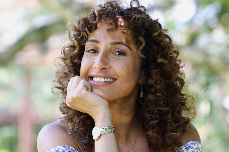 Uma mulher com cabelos cacheados e castanhos, usando um vestido de alças com estampa floral. Ela está sentada em um ambiente ao ar livre, sorrindo e apoiando o queixo na mão. Ao fundo, há árvores e um ambiente natural desfocado.