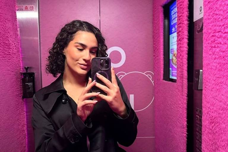 A imagem mostra uma pessoa tirando uma selfie em um elevador com paredes rosa. A pessoa tem cabelo cacheado e usa um casaco preto. Ao fundo, há um painel com telas e um design simples. O ambiente é bem iluminado e moderno.