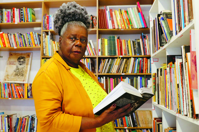 Uma mulher de pele escura e cabelo grisalho, preso em um coque, está em uma biblioteca segurando um livro aberto. Ela usa uma blusa amarela com bolinhas pretas e um casaco laranja. Ao fundo, há prateleiras repletas de livros de várias cores e tamanhos.