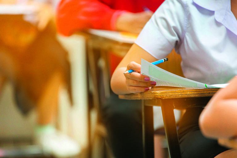 A imagem mostra uma mão de um estudante segurando um lápis e uma folha de papel em uma mesa de escola. Ao fundo, é possível ver outros alunos, mas eles estão desfocados. A cena sugere um ambiente de sala de aula durante uma prova.