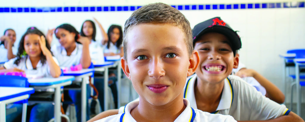 A imagem mostra um grupo de crianças em uma sala de aula. Em primeiro plano, um menino de cabelo curto e liso, com uma camiseta branca e gola azul, está sorrindo para a câmera. Atrás dele, um outro menino, também sorridente e usando um boné preto, faz uma expressão divertida. Ao fundo, várias meninas e meninos estão sentados em mesas, alguns com as mãos levantadas, parecendo se divertir.