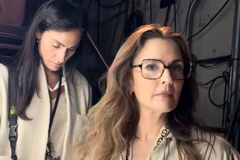 A imagem mostra duas mulheres em um ambiente de bastidores. A mulher à frente tem cabelos longos e ondulados, usa óculos e uma camisa listrada. A mulher ao fundo está vestindo um casaco branco e parece estar organizando algo. O ambiente é levemente iluminado e há fios e equipamentos visíveis ao fundo.