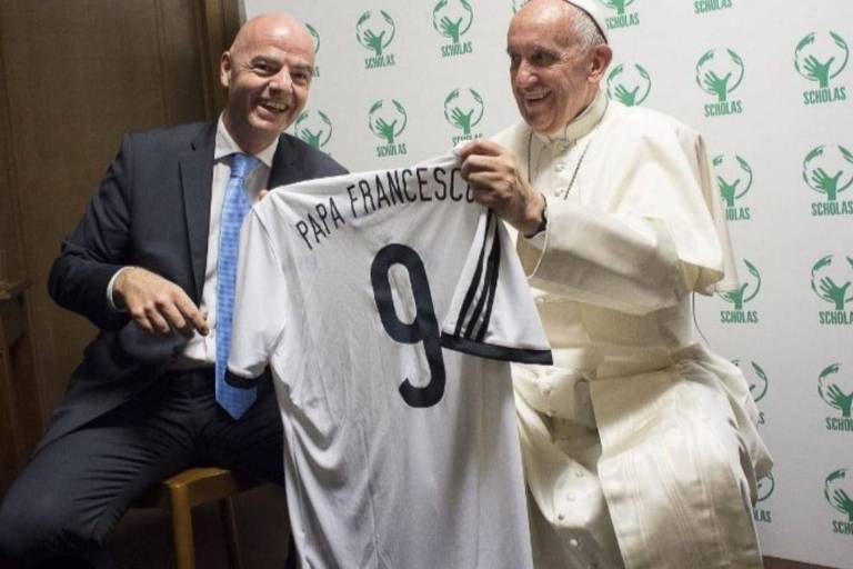 Na imagem, o Papa Francisco está segurando uma camisa de futebol branca com o nome "PAPA FRANCESCO" e o número 9. Ao seu lado, um homem sorridente, vestido com um terno escuro e gravata, também segura a camisa. O fundo é decorado com um padrão verde que inclui o logotipo de uma organização chamada "Scholas".