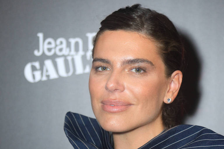 A imagem mostra uma mulher em um evento de moda, posando para a câmera. Ela está usando um vestido com listras verticais e um detalhe de laço na parte superior. O fundo é escuro, com o nome 'Jean Paul Gaultier' visível em letras brancas. A mulher tem cabelo preso e está sorrindo levemente.