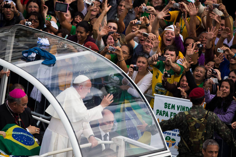 Vestido de branco, um homem acena dentro de um carro com parede de vidro e laterais abertais para uma aglomeração de jovens, muito com camisas amarelas e bandeiras do Brasil