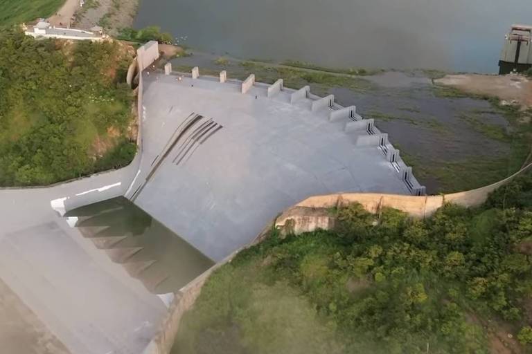 A imagem mostra uma vista aérea de uma barragem, com uma estrutura de concreto que se estende até a água. A barragem é acompanhada por um grande corpo d'água, com várias ilhas visíveis ao fundo. A vegetação verdejante está presente nas margens, e o céu está parcialmente nublado.