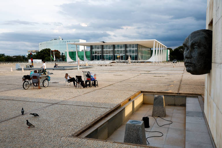 A imagem mostra uma praça ampla com um piso de pedras. No primeiro plano, há uma escultura de um rosto humano em relevo na parede à direita (busto de JK). Algumas pessoas estão sentadas em cadeiras e bancos. No fundo, está o prédio do STF, um edifício moderno com grandes janelas e estrutura arquitetônica distinta