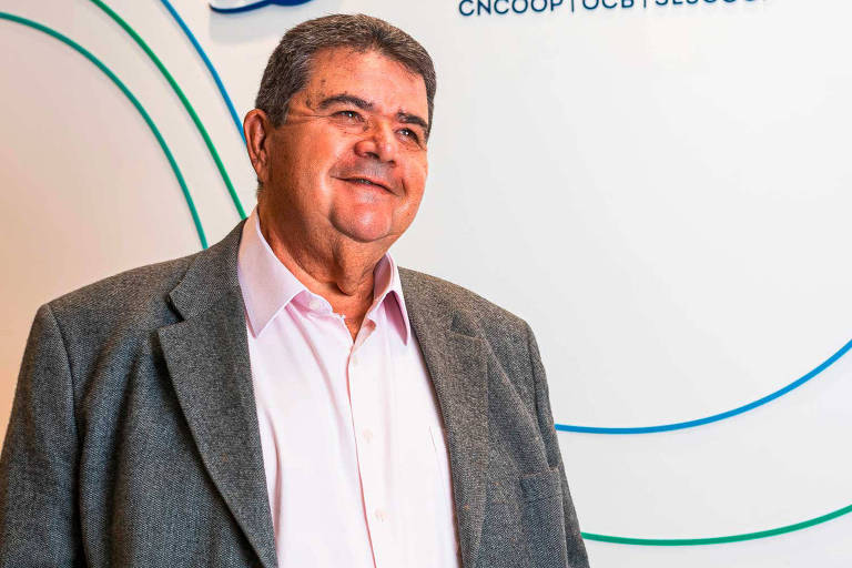 Um homem sorridente está posando para a foto. Ele usa um paletó cinza e uma camisa rosa. O fundo é claro, com linhas coloridas em verde e azul, e contém texto que parece ser relacionado a uma organização ou evento. O homem parece estar em um ambiente profissional.