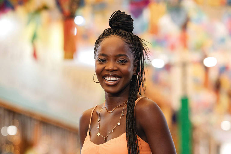 Uma mulher sorridente está em um ambiente interno decorado com bandeirinhas coloridas. Ela usa um macacão laranja e segura uma bolsa de palha. O fundo apresenta uma variedade de itens, sugerindo um mercado ou feira. A iluminação é suave e acolhedora, destacando a alegria da mulher.