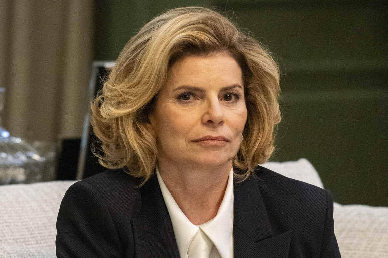 Uma mulher está sentada em um sofá claro, vestindo um blazer preto sobre uma camisa branca e calças brancas. Ela tem cabelo loiro e ondulado, e está olhando para a frente com uma expressão neutra. O ambiente ao redor é moderno, com plantas e objetos decorativos visíveis ao fundo.