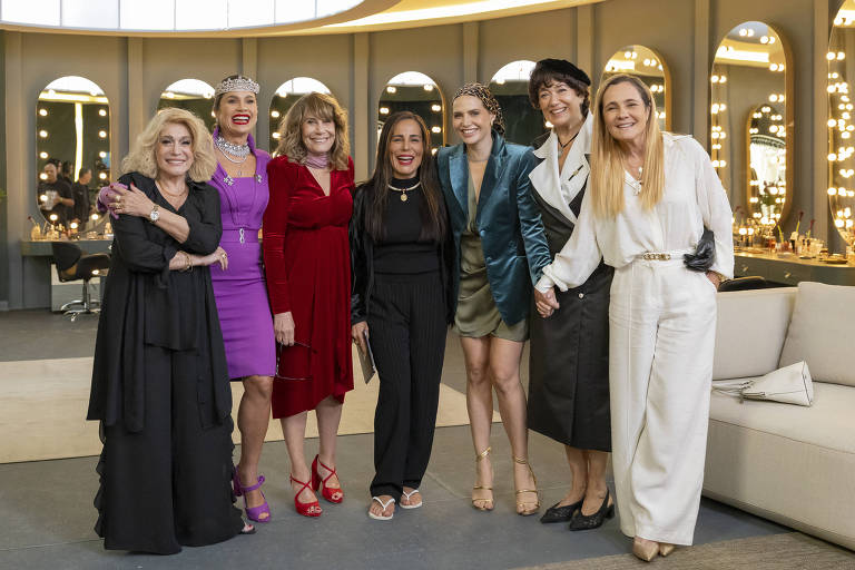Um grupo de oito mulheres posando juntas em um ambiente bem iluminado, com espelhos e luzes ao fundo. Elas estão vestidas de forma elegante, algumas com vestidos coloridos e outras com roupas mais formais. Todas estão sorrindo e parecem estar se divertindo. O ambiente é moderno e sofisticado, com um sofá claro ao lado.