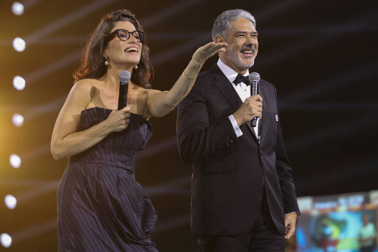 Dois apresentadores estão em um palco iluminado, sorrindo e segurando microfones. A mulher, com cabelo solto e óculos, usa um vestido escuro e gesticula enquanto fala. O homem, com cabelo grisalho e terno escuro, sorri e olha para o público. Ao fundo, há luzes e uma tela com imagens.