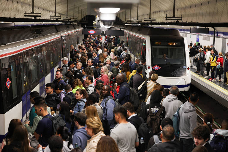 A imagem mostra uma estação de metrô lotada com muitas pessoas aguardando para embarcar. Dois trens estão visíveis, um deles está parado na plataforma enquanto o outro está parcialmente visível. A multidão é diversificada, com pessoas de diferentes idades e etnias. O ambiente é interno, com iluminação artificial e paredes de azulejos.