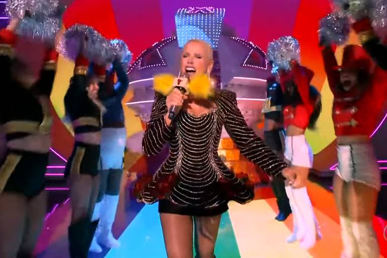 Uma artista está se apresentando em um palco colorido, segurando um microfone. Ela usa um vestido preto com detalhes brilhantes e uma saia volumosa. Atrás dela, dançarinos estão levantando pompons prateados, vestindo trajes de gala em cores variadas. O fundo do palco é iluminado com luzes em um padrão de arco-íris, criando um ambiente vibrante e festivo.