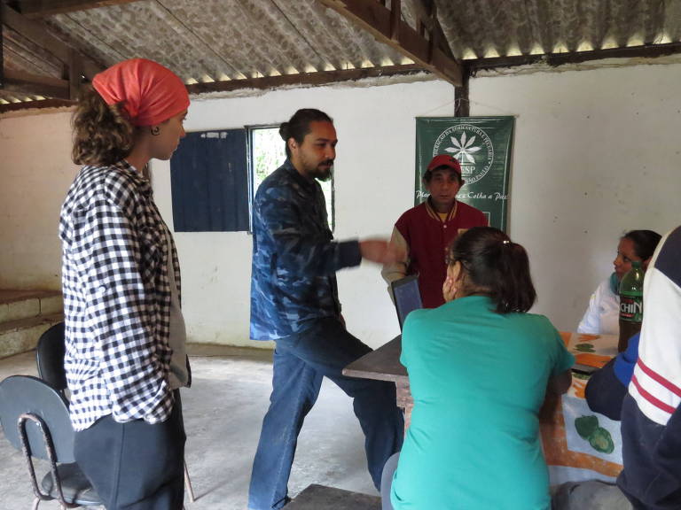 A imagem mostra um grupo de pessoas em um ambiente interno, possivelmente uma sala de reuniões. Um homem com cabelo longo e barba, vestido com uma camisa azul, está em pé e gesticulando enquanto fala. Uma mulher com um lenço vermelho na cabeça observa. Outras pessoas estão sentadas ao redor de uma mesa, algumas delas de costas para a câmera. Ao fundo, há uma bandeira verde com um símbolo, e uma janela aberta.