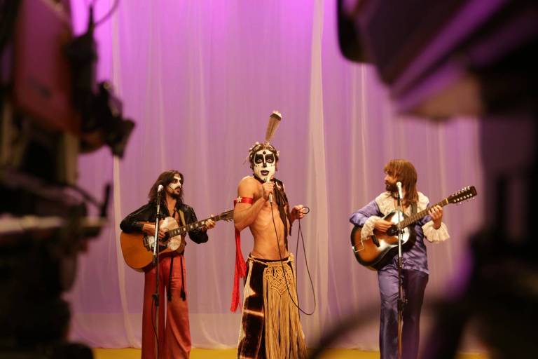 A imagem mostra três músicos se apresentando em um palco. O músico central está vestido com uma roupa exótica e pintado, segurando um microfone. Os outros dois músicos estão à sua esquerda e direita, tocando violões. O fundo é iluminado com luzes roxas e brancas, e há uma câmera visível em primeiro plano, sugerindo que a apresentação está sendo gravada.