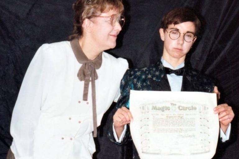 A imagem mostra duas pessoas em um evento de premiação. À esquerda, uma mulher com cabelo castanho claro e óculos, vestindo uma blusa branca com um laço. À direita, um homem com cabelo curto e óculos, usando um terno preto com uma camisa branca e gravata. Ele segura um diploma ou certificado que diz 'Magia Circo'. O fundo da imagem é preto.