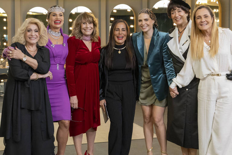 Um grupo de oito mulheres posando juntas em um ambiente bem iluminado, com espelhos e luzes ao fundo. Elas estão vestidas de forma elegante, algumas com vestidos coloridos e outras com roupas mais formais. Todas estão sorrindo e parecem estar se divertindo. O ambiente é moderno e sofisticado, com um sofá claro ao lado.