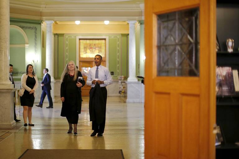 A imagem mostra um interior de um edifício público, com paredes em tom verde e detalhes em mármore. No centro, há um grupo de pessoas, incluindo um homem sorridente vestido com uma camisa branca e calças escuras, e uma mulher com cabelo longo e cinza. Outros indivíduos estão visíveis ao fundo, alguns em trajes formais. Uma porta de madeira está parcialmente aberta, revelando o interior do espaço. O ambiente é bem iluminado e decorado.