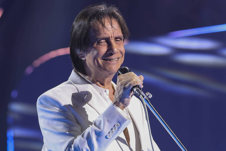 Um cantor está se apresentando em um palco iluminado. Ele usa um terno branco e segura um microfone com um cabo. O fundo é composto por luzes coloridas e formas circulares, criando um ambiente vibrante e dinâmico.