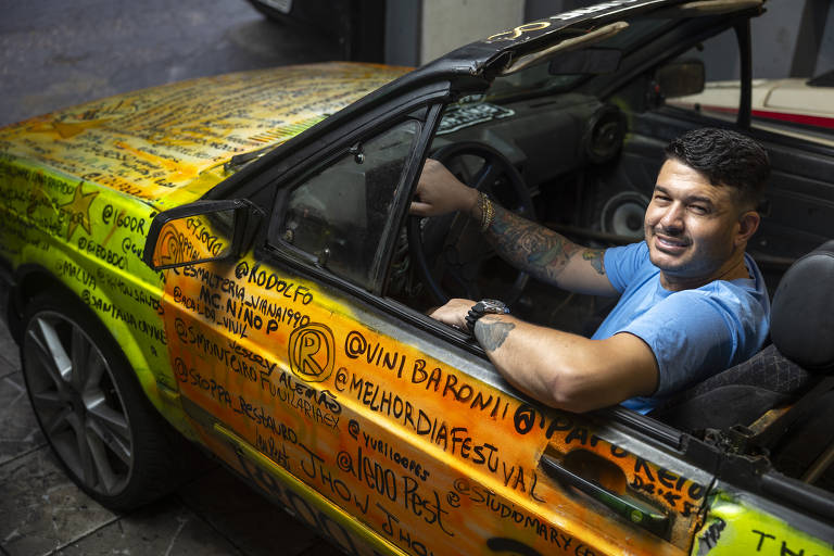 Um homem está sentado no interior de um carro esportivo, que possui uma pintura colorida com várias mensagens e nomes escritos em tinta preta e laranja. O homem sorri e usa uma camiseta azul, enquanto exibe tatuagens nos braços. O carro está estacionado em um ambiente interno, com luz natural iluminando a cena.