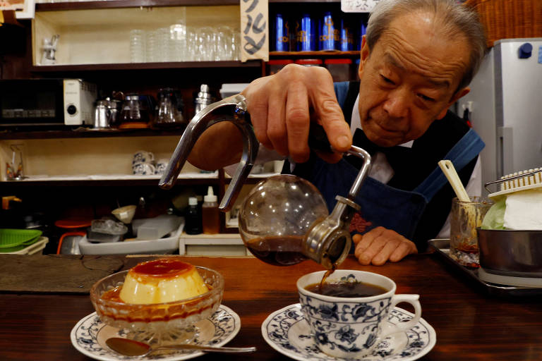 Um homem idoso está servindo café de uma jarra de vidro em uma xícara de porcelana. Ao lado dele, há uma sobremesa de pudim com calda de caramelo em um prato decorativo. O ambiente é acolhedor, com prateleiras ao fundo contendo utensílios de café e outros itens de cozinha.