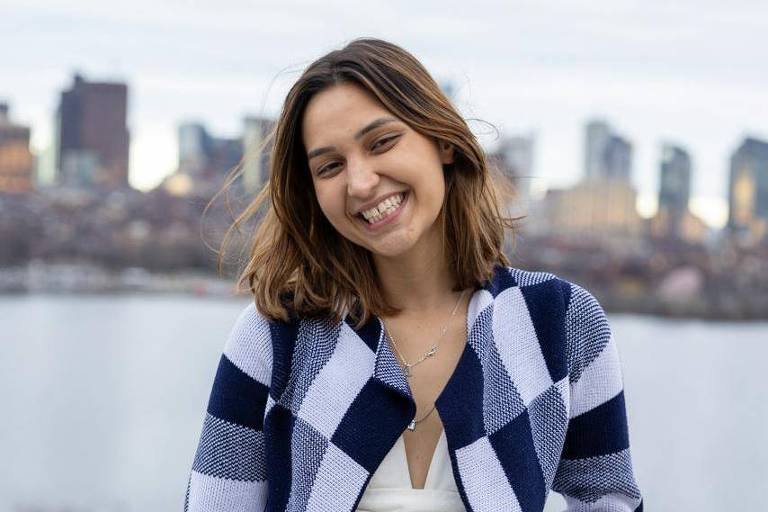 Uma mulher está sorrindo enquanto posa para a foto em uma varanda. Ela usa um casaco listrado em azul e branco e uma blusa branca. Ao fundo, há uma vista de uma cidade com prédios e um corpo d'água. O céu está nublado, e a cena transmite uma sensação de tranquilidade.