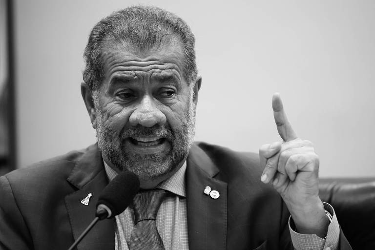 Um homem com barba e cabelo grisalho, vestindo um terno azul e uma gravata, está falando em um microfone. Ele levanta um dedo, como se estivesse enfatizando um ponto durante sua fala. O fundo é desfocado, mas parece ser um ambiente de reunião ou conferência.