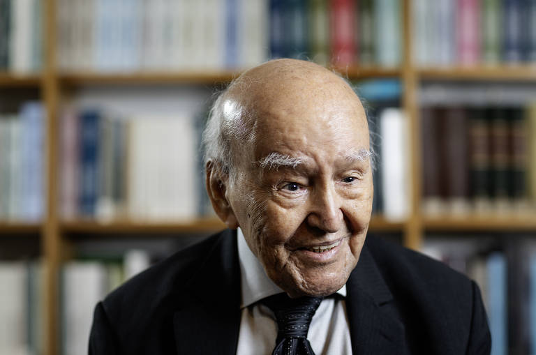 Um homem idoso está sorrindo, vestindo um terno escuro e uma gravata. Ele tem cabelo grisalho e uma expressão amigável. Ao fundo, há uma estante com livros organizados, sugerindo um ambiente de estudo ou biblioteca.