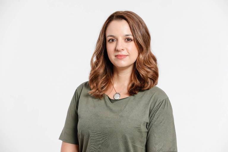A imagem mostra uma mulher com cabelo castanho claro e ondulado, usando uma blusa verde. Ela está de pé, com um leve sorriso, olhando diretamente para a câmera. O fundo é branco e neutro, destacando a figura da mulher.