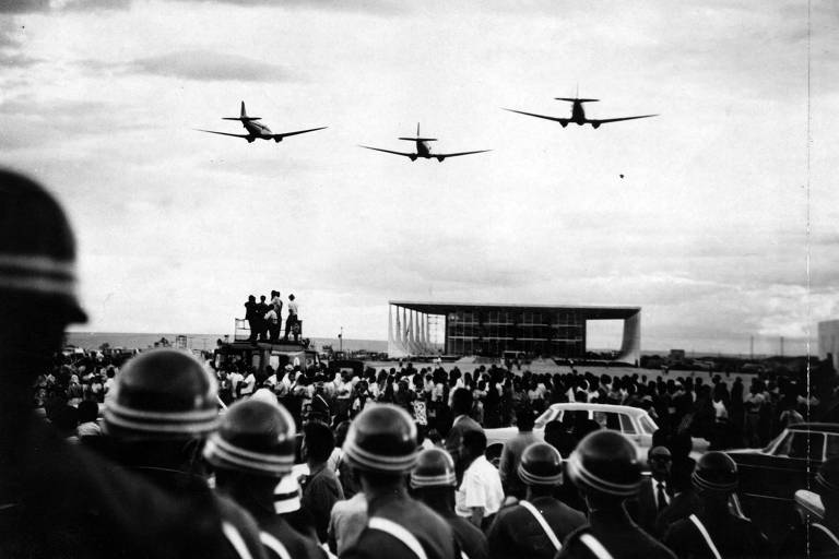 A imagem mostra uma multidão reunida em um evento ao ar livre, com várias pessoas em pé e algumas em um caminhão. No céu, três aviões estão voando em formação. Em primeiro plano, há um grupo de soldados com capacetes, observando a cena. Ao fundo, um edifício moderno é visível, e há carros estacionados na área