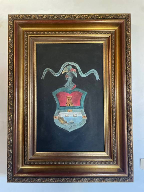 A imagem mostra um escudo emoldurado com uma moldura dourada elaborada. O escudo apresenta uma figura central com um fundo escuro, onde se destaca uma figura humana estilizada com um chapéu ou elmo, adornado com uma fita. O escudo tem cores predominantes em vermelho e azul, com detalhes em branco e dourado.