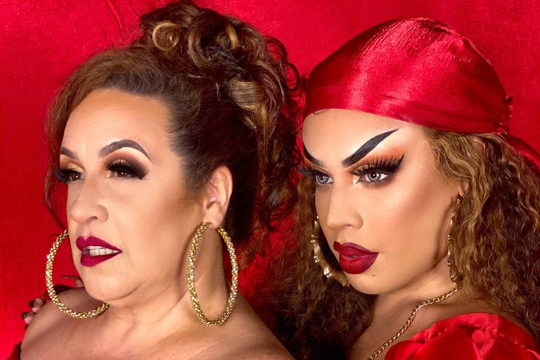 Imagem mostra duas mulheres com roupas vermelhas e batom vermelho. Ambas são brancas e posam para a foto frente a um tecido vermelho
