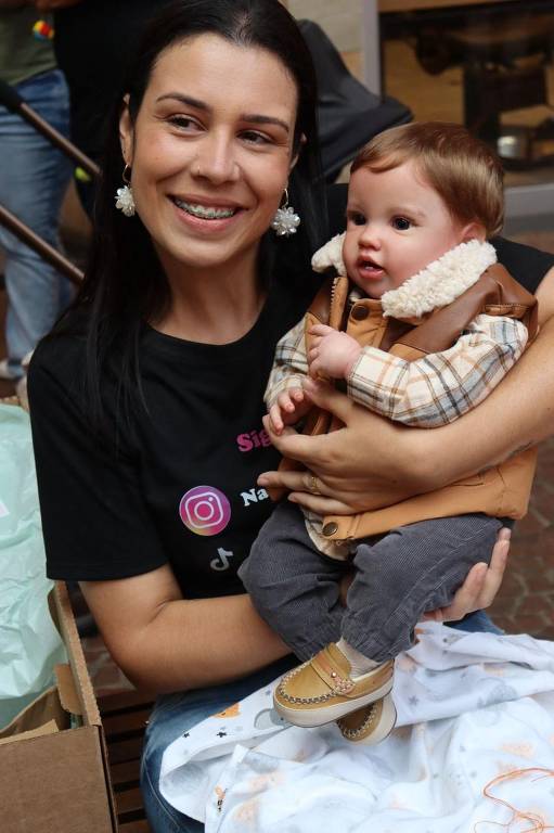 Uma mulher sorridente está segurando um boneco que se parece com um bebê. Ela usa uma camiseta preta e brincos grandes. O boneco está vestido com um colete marrom e uma camisa xadrez. Ao fundo, há pessoas e uma caixa com papel de presente. A mulher parece estar em um ambiente festivo.
