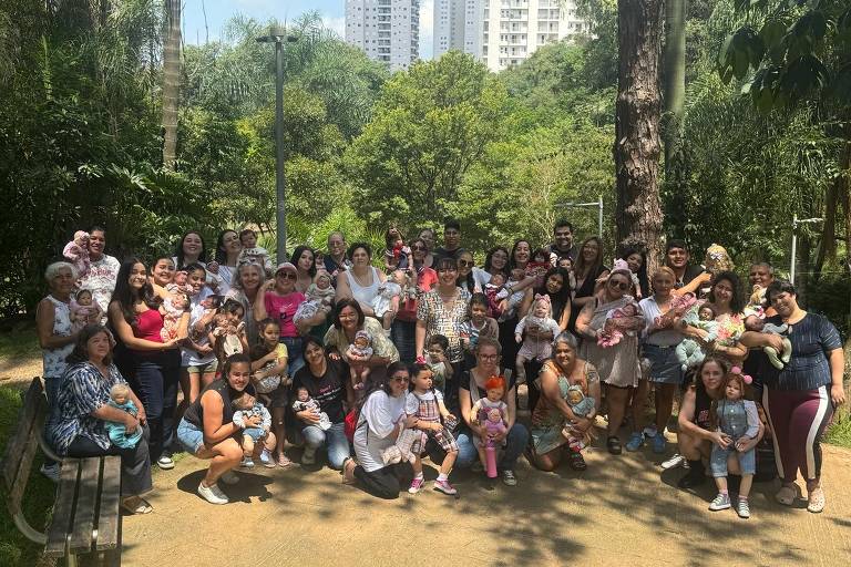A imagem mostra um grande grupo de pessoas reunidas em um parque. Há muitas mulheres, algumas com bebês nos braços, e algumas crianças. O cenário é verde, com árvores e prédios ao fundo. As pessoas estão sorrindo e posando para a foto, criando um ambiente alegre e comunitário.