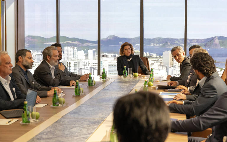 A imagem mostra uma reunião em uma sala de conferência moderna, com uma longa mesa retangular. Há várias pessoas sentadas ao redor da mesa, algumas com laptops e documentos. No fundo, grandes janelas oferecem uma vista panorâmica da cidade e das montanhas ao longe. A iluminação é natural, e a atmosfera parece ser de discussão séria.