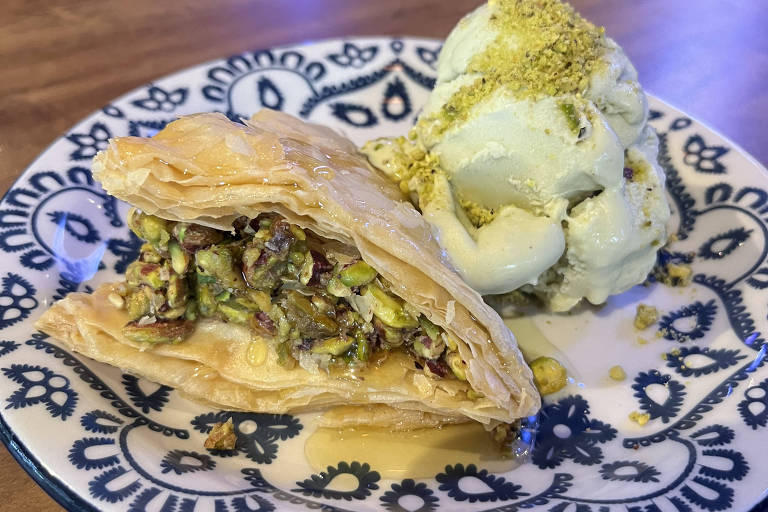 Um prato com uma porção de baklava, que é um doce folhado recheado com pistaches e nozes, ao lado de uma bola de sorvete de pistache, polvilhada com pistaches picados. O prato é decorado com um padrão azul e branco.