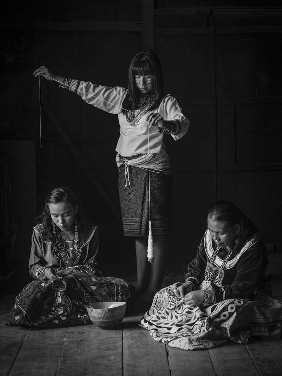 A imagem em preto e branco mostra três mulheres em um ambiente interno. Uma mulher está em pé, segurando um fio, enquanto as outras duas estão sentadas no chão, uma delas está bordando e a outra observa. Todas estão vestidas com roupas tradicionais. O ambiente parece rústico, com paredes de madeira ao fundo.